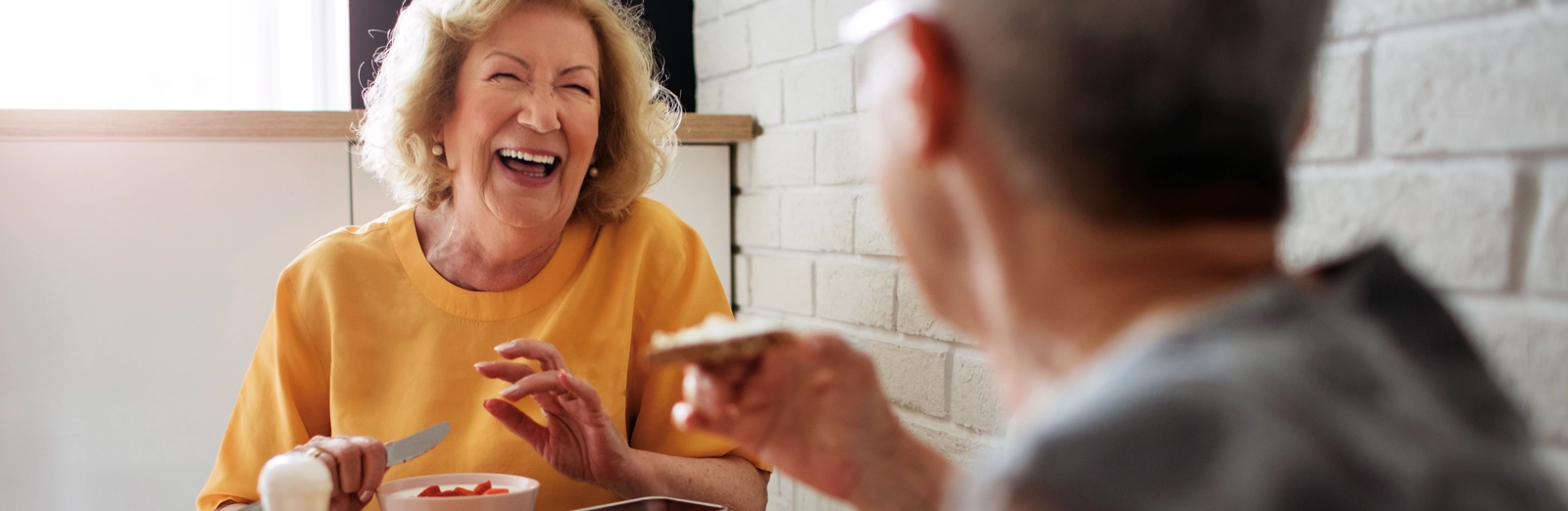 older-couple-laughing-while-eating-breakfast-1600x522.webp